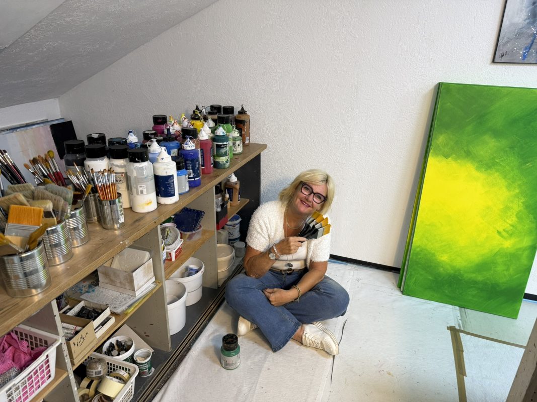 Künstlerin Elisabeth Suter sitzend mit Pinseln neben einem grünen Originalkunstwerk im Atelier